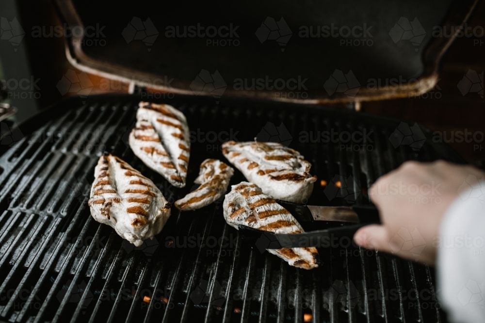 Grilled chicken breast on a BBQ being turned by a hand holding tongs - Australian Stock Image