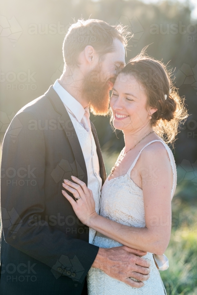 Groom gives bride a kiss against the sun light rays on their wedding day. - Australian Stock Image