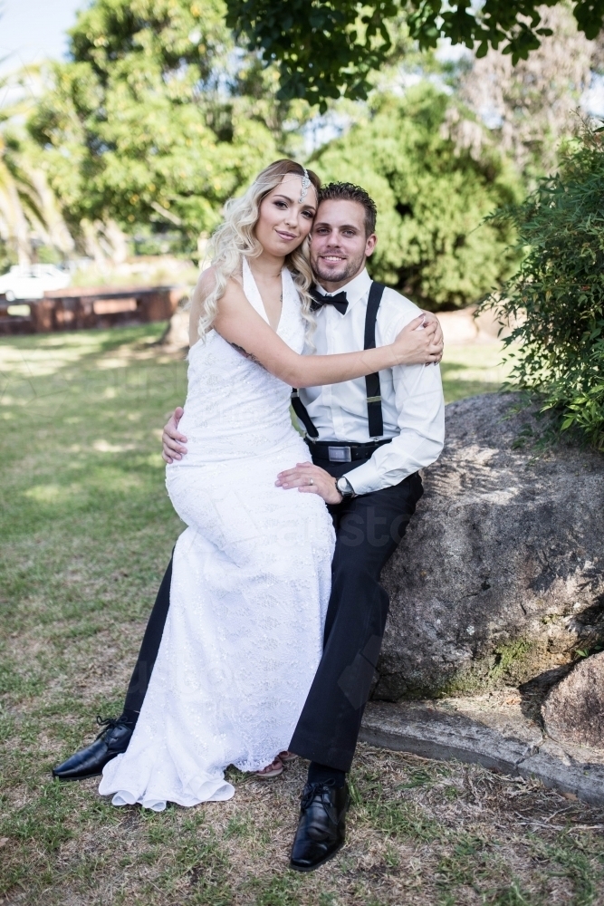 Groom sitting on rock with bride on his lap in garden - Australian Stock Image