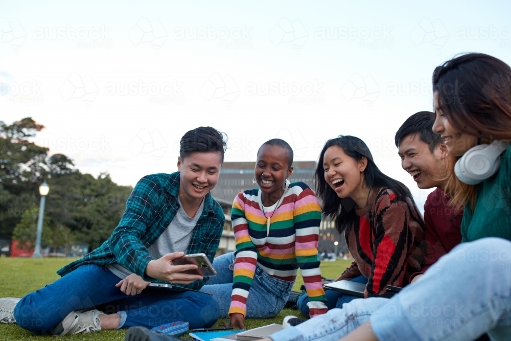 Group of young university students hanging out sitting on grass studying and using devices - Australian Stock Image