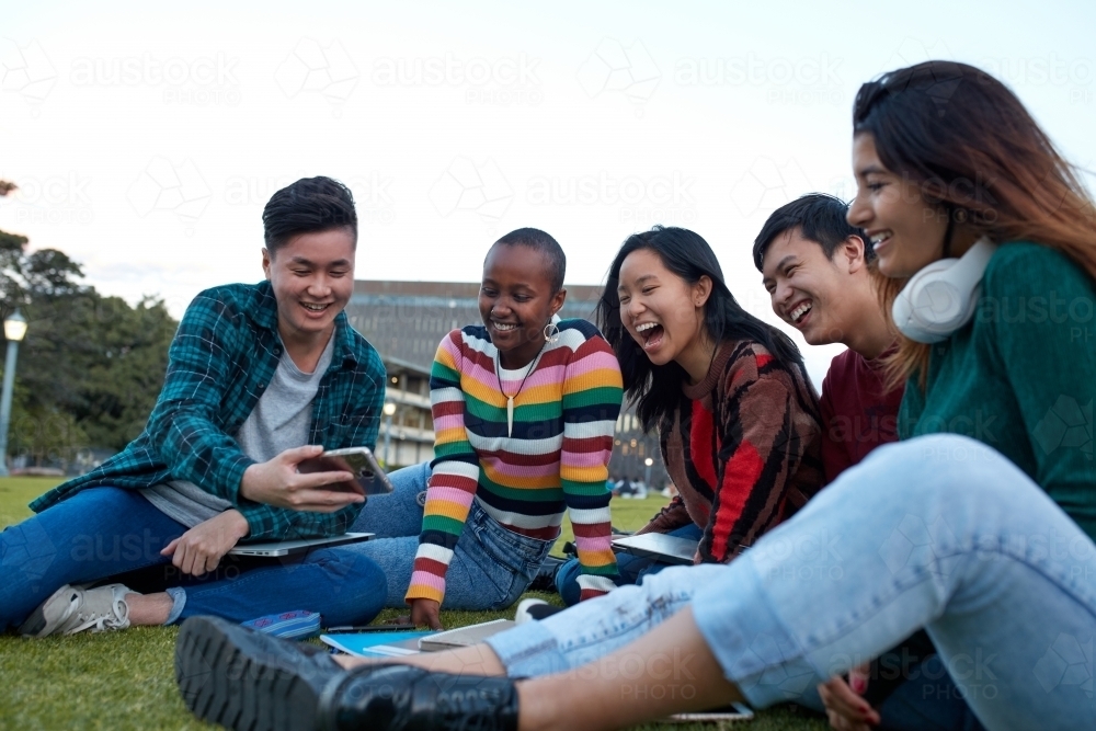 Group of young university students hanging out sitting on grass studying and using devices - Australian Stock Image