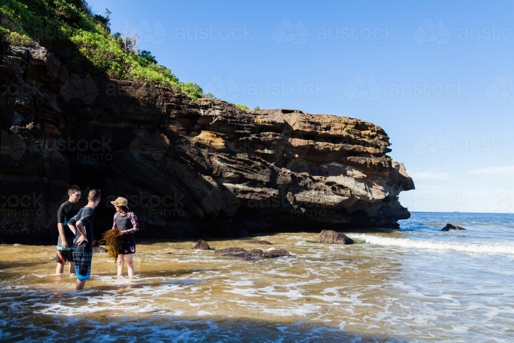 Group of youth standing in the ocean beside sea cliff on sunny day - Australian Stock Image