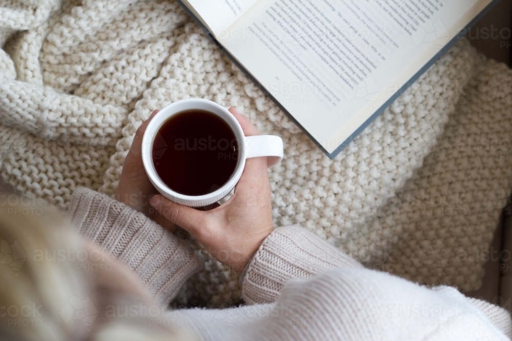 Hands holding coffee mug with woollen blanket and book - Australian Stock Image