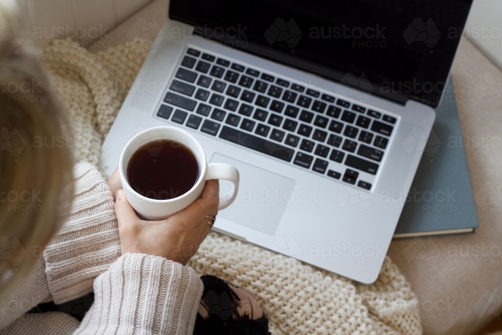 Hands holding coffee mug with woollen blanket and laptop computer and book - Australian Stock Image