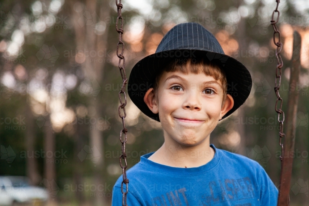 Happy 9yo kid in a hat playing on swing outside - Australian Stock Image