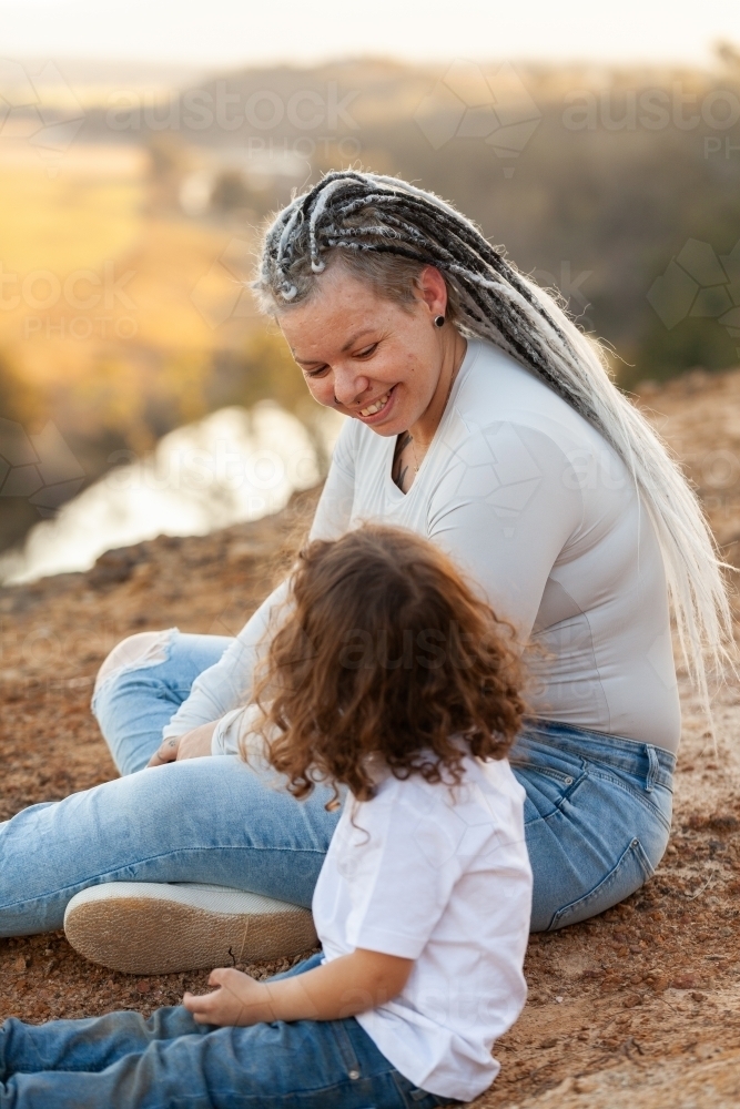 Happy aboriginal mum with daughter sitting on cliff edge in country telling stories - Australian Stock Image