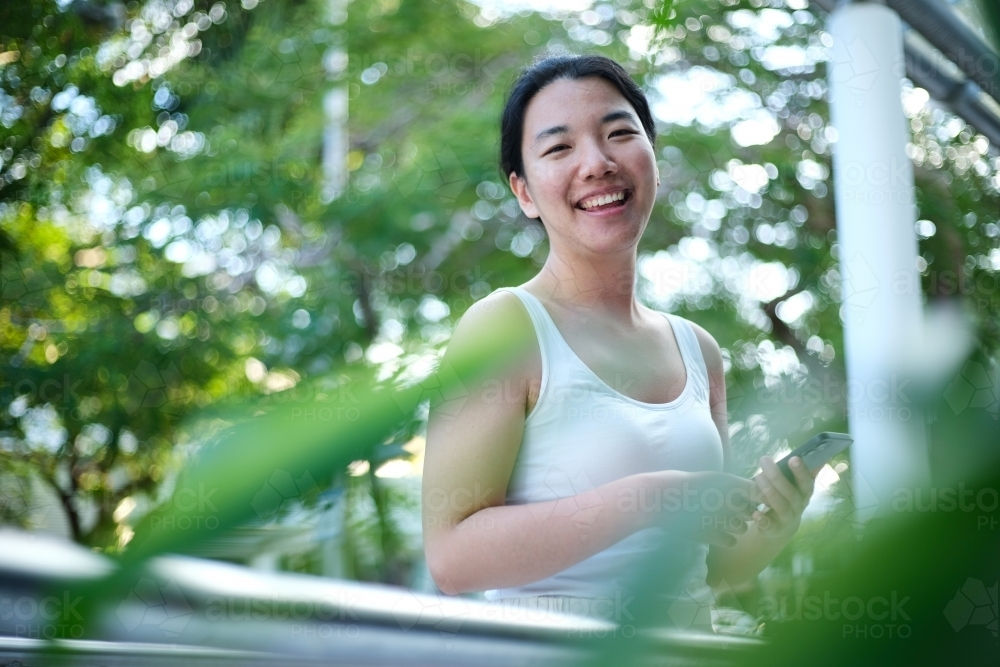 Happy Asian woman standing along walkway outdoors with green bokeh backdrop - Australian Stock Image