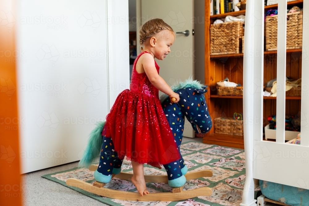 Happy Aussie kid in red dress riding on rocking horse toy in her bedroom - Australian Stock Image