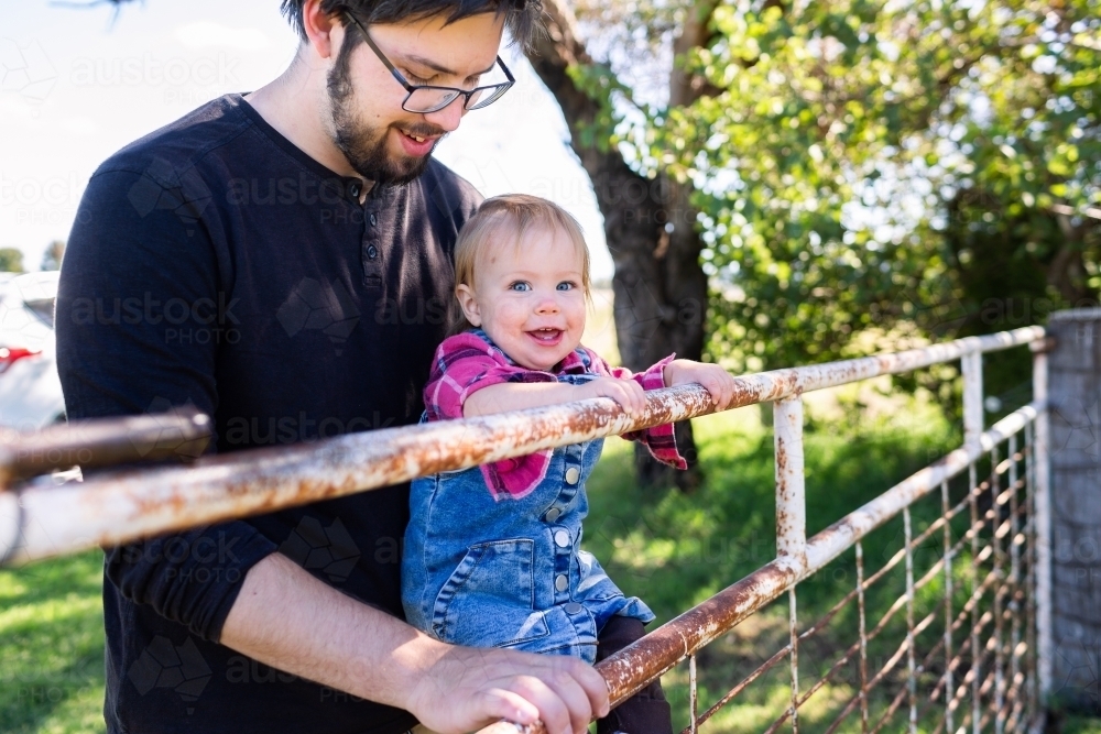 Happy baby country kid swinging on farm gate with dad - Australian Stock Image