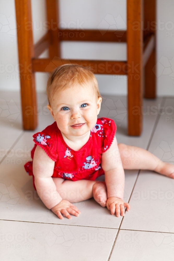 Happy baby girl in red Christmassy outfit sitting on kitchen floor - Australian Stock Image