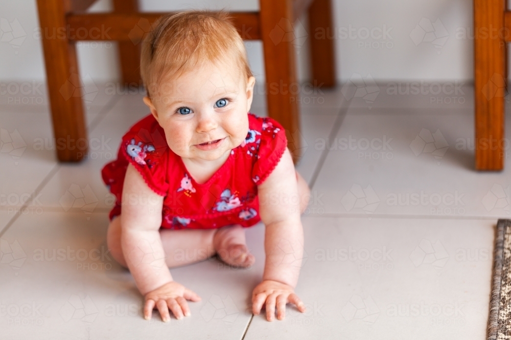 Happy baby girl in red Christmassy outfit sitting on kitchen floor - Australian Stock Image