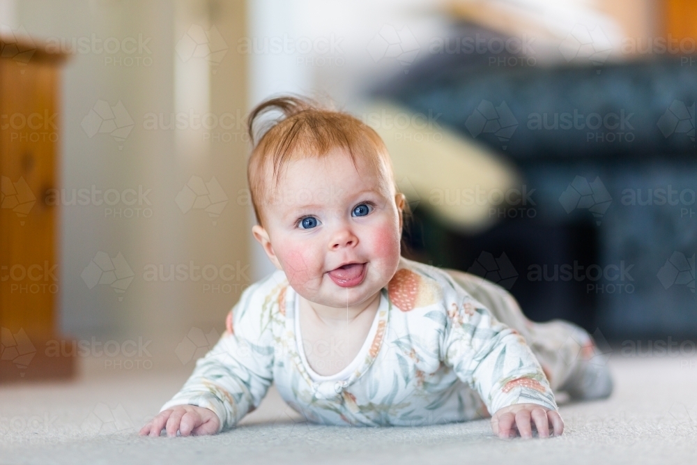 Happy baby girl learning to crawl doing tummy time on living room floor - Australian Stock Image