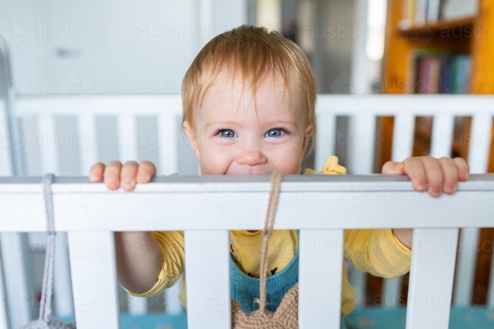 happy baby girl peering over cot bars in bedroom - Australian Stock Image