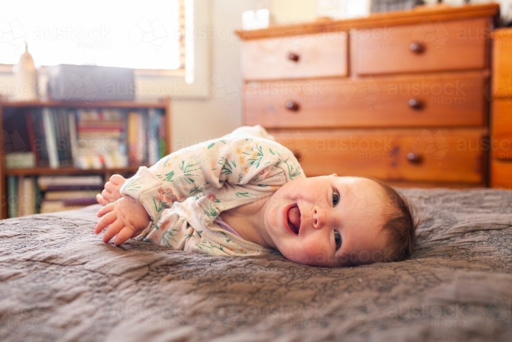 Happy baby girl trying to roll over on parents bed in bedroom - Australian Stock Image