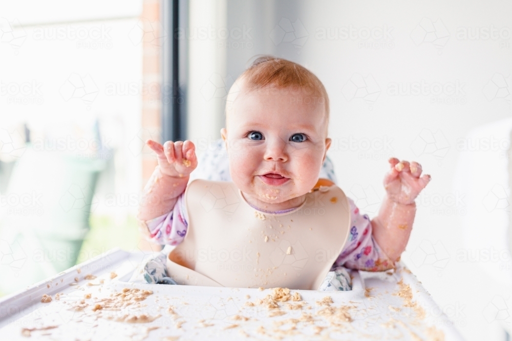 Happy baby in high chair with messy food - Australian Stock Image