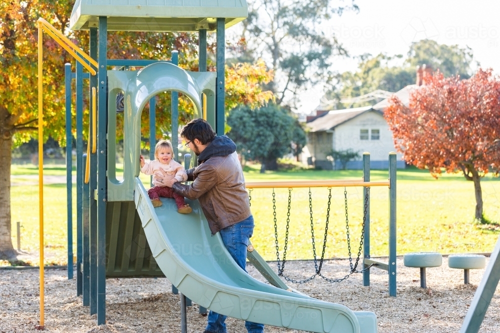 Happy baby playing at park with father sliding down slippery dip - Australian Stock Image