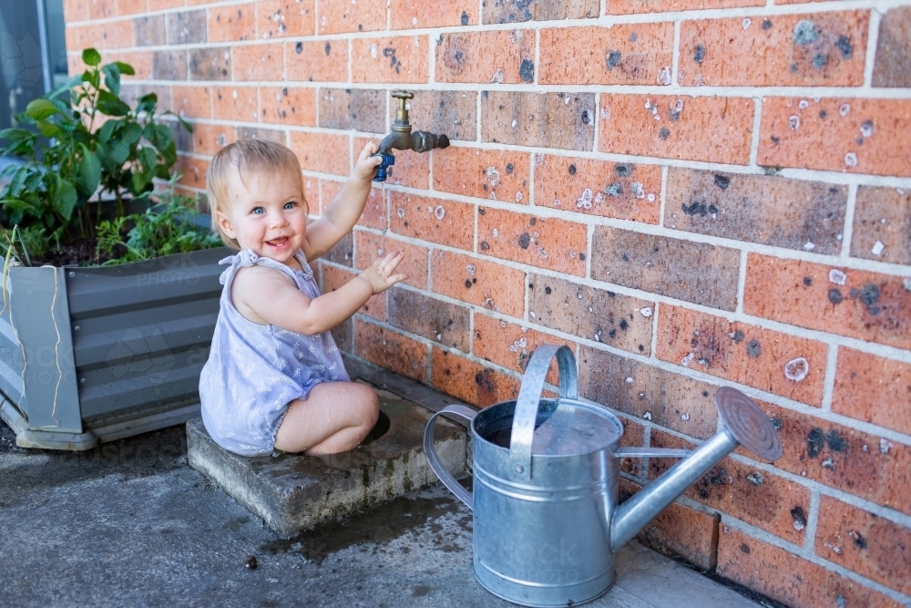 Happy baby smiling sitting in tap water drain in backyard with watering can - Australian Stock Image