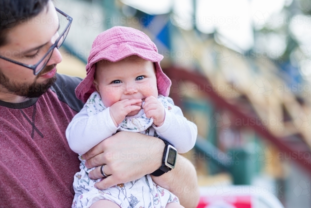 Happy baby wearing hat outside in dads arms with grandstand behind at showground - Australian Stock Image