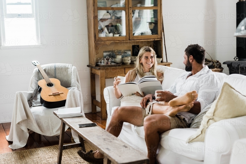 Happy couple relaxing on a couch talking, reading and drinking coffee - Australian Stock Image