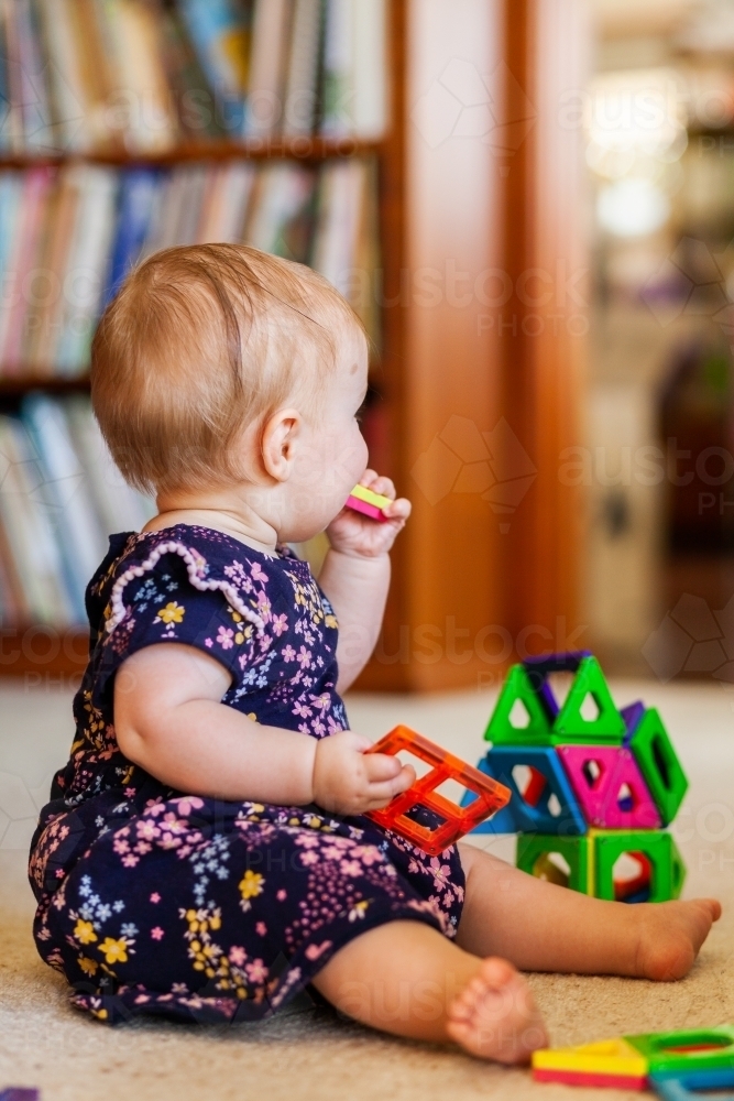 Happy eight month old baby sitting on floor playing with magnetic blocks - Australian Stock Image