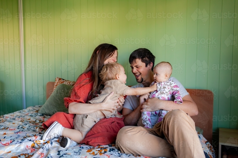 Happy family of four sitting together on bed in home with toddler and baby laughing - Australian Stock Image