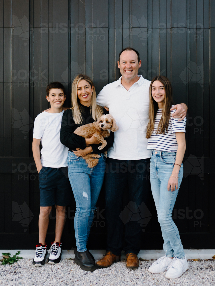 Happy family standing outside in front of black garage door with pet dog - Australian Stock Image