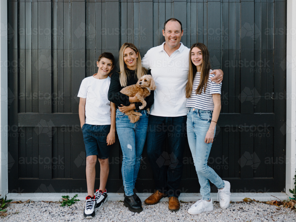 Happy family standing outside in front of black garage door with pet dog - Australian Stock Image