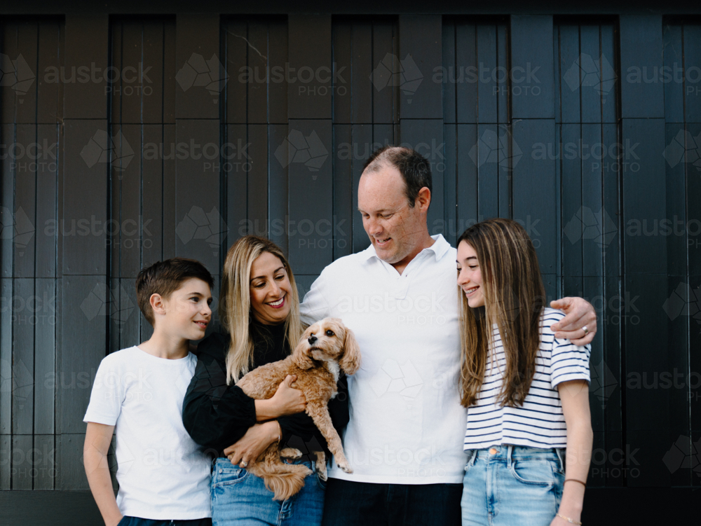 Happy family standing outside in front of black garage door with pet dog - Australian Stock Image