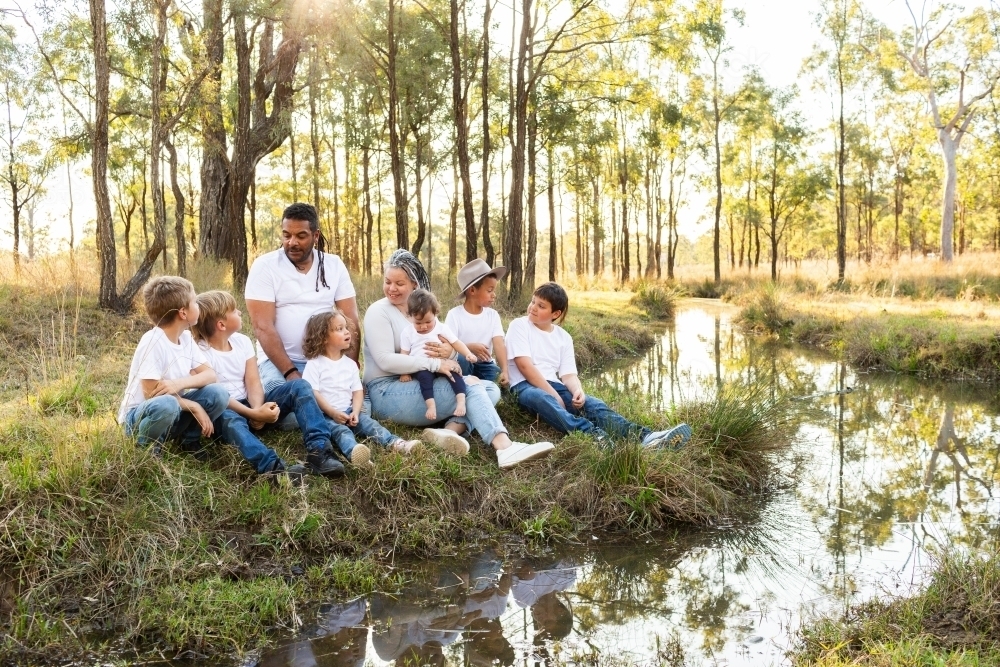 Happy First nations Australian family sitting together by creek in rural paddock - Australian Stock Image