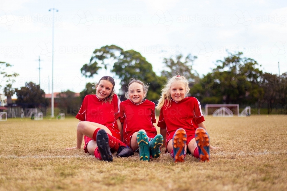 Happy girls wearing red soccer uniforms sitting on the grass together - Australian Stock Image