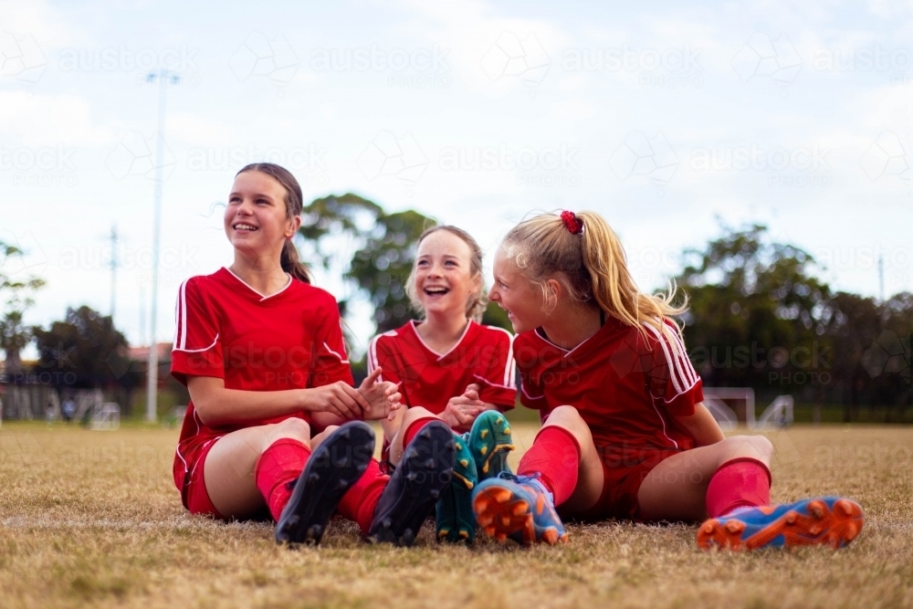 Happy girls wearing red soccer uniforms sitting on the grass together - Australian Stock Image