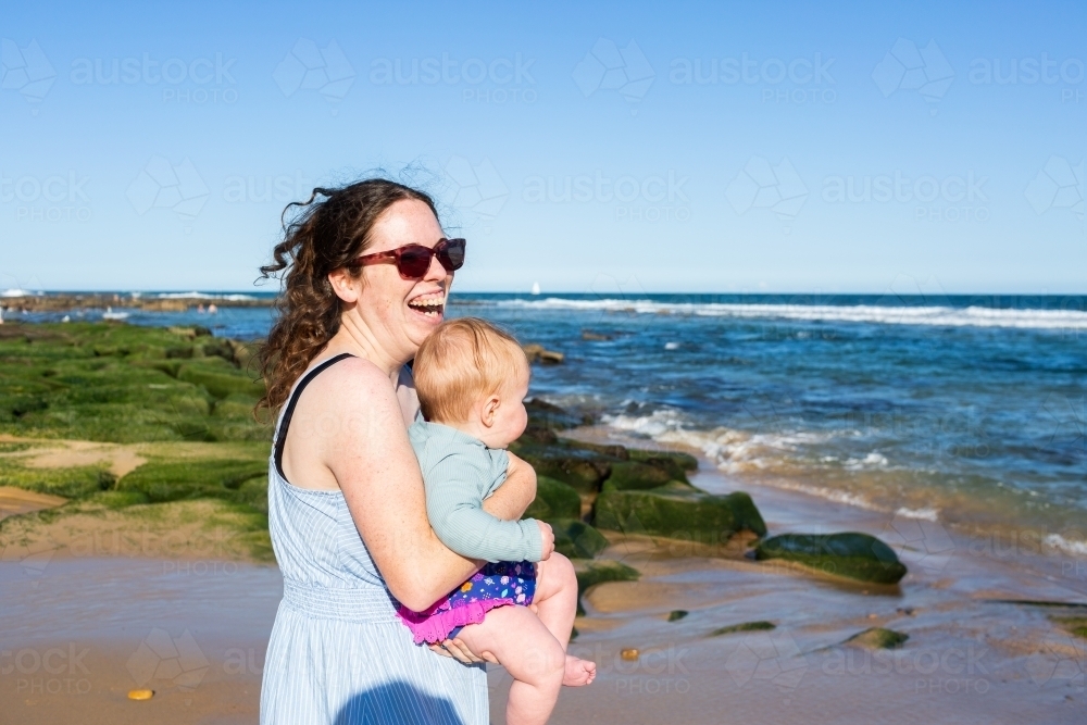 happy laughing mother carrying baby girl towards ocean at beach - Australian Stock Image