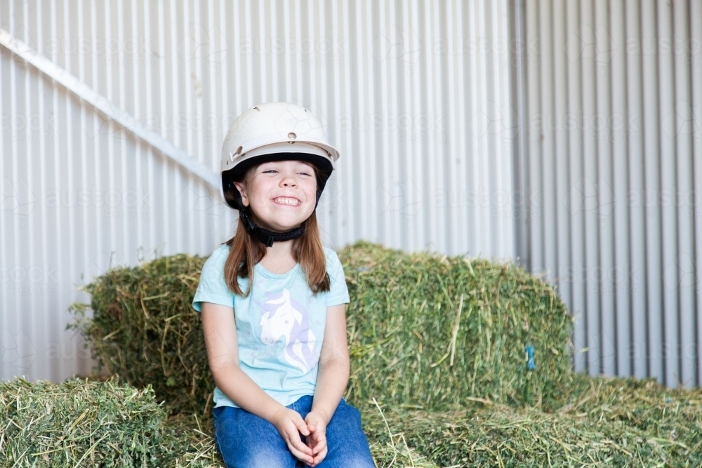 Happy little girl in horse riding helmet sitting on lucerne hay bales - Australian Stock Image