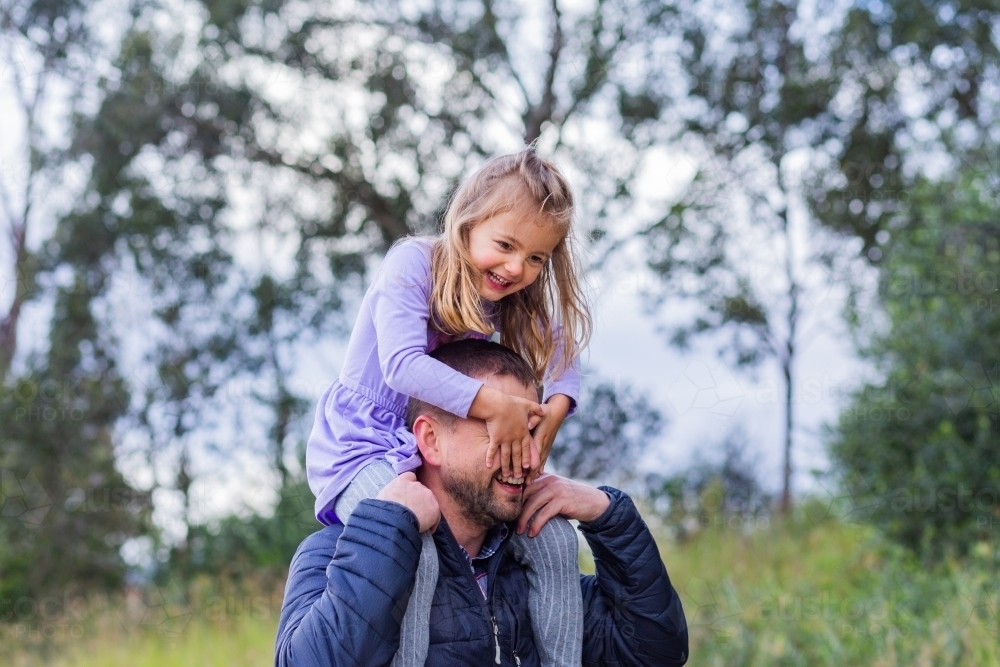 Happy little girl riding on her dads shoulders covering his face - Australian Stock Image