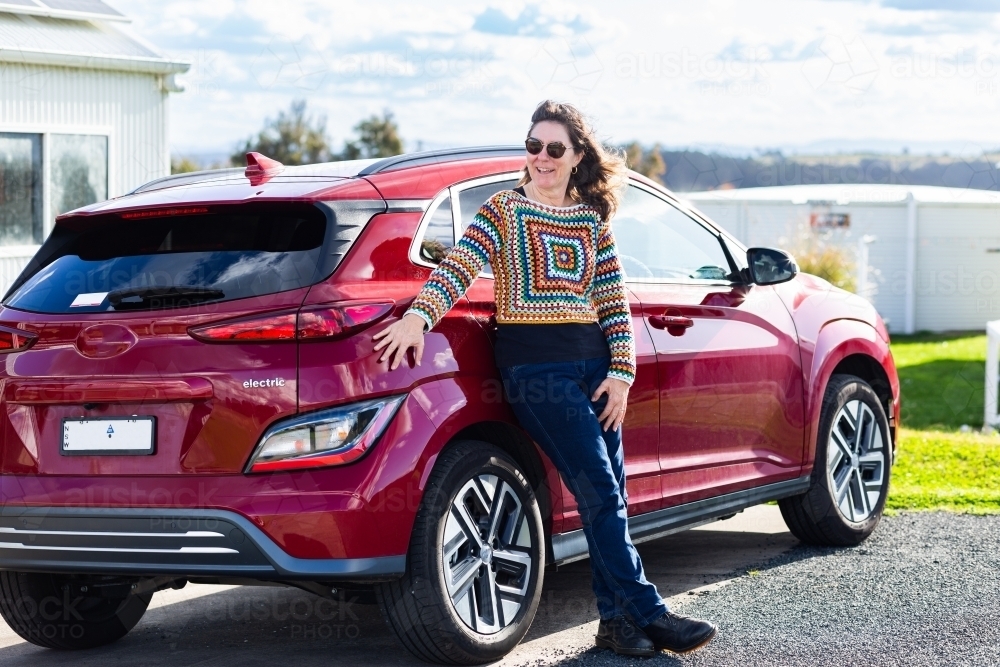 happy middle age woman standing beside electric vehicle car - Australian Stock Image