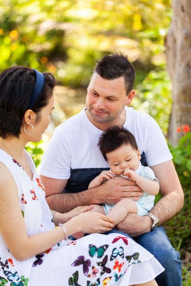 happy mixed race family of three siting together outside in garden - Australian Stock Image