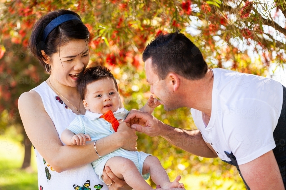 Happy mixed race family of three with four month old baby laughing together outside - Australian Stock Image