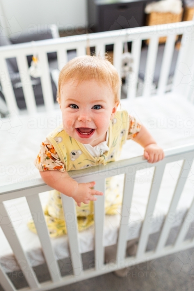 Happy nine month old baby standing up in cot smiling in delight in morning - Australian Stock Image