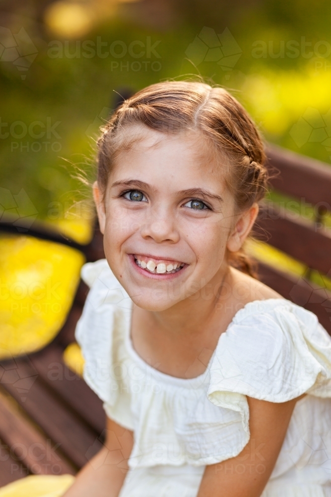 Happy nine year old girl in yellow dress sitting on park bench - Australian Stock Image