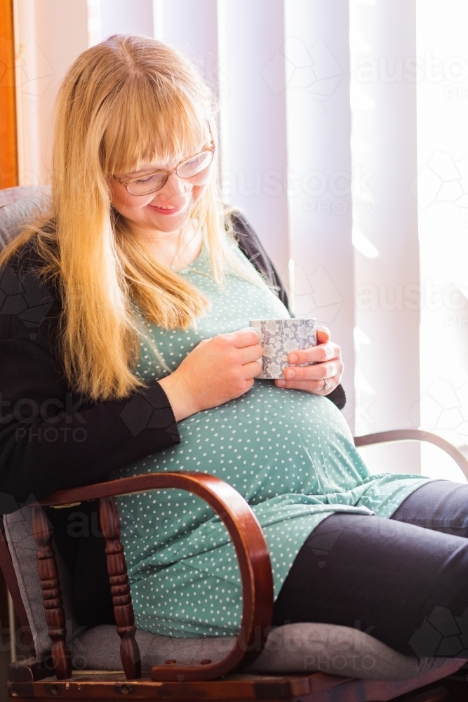 Happy pregnant woman sitting in rocking chair with mug of coffee - Australian Stock Image