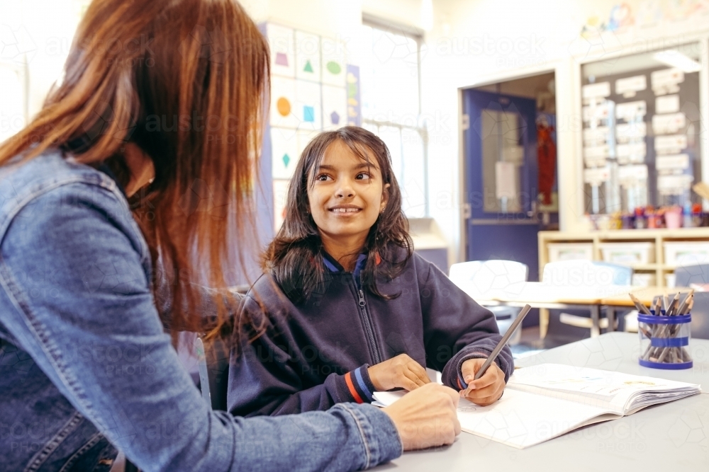 Happy primary school student listening to her teacher whilst writing - Australian Stock Image