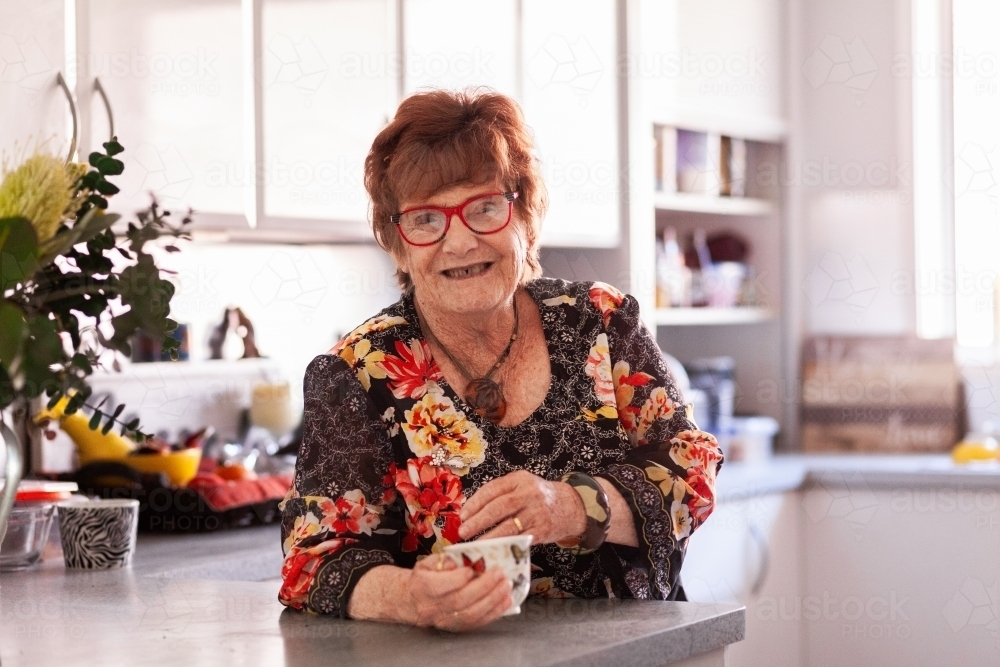 Happy senior woman in her kitchen drinking tea from cup - Australian Stock Image