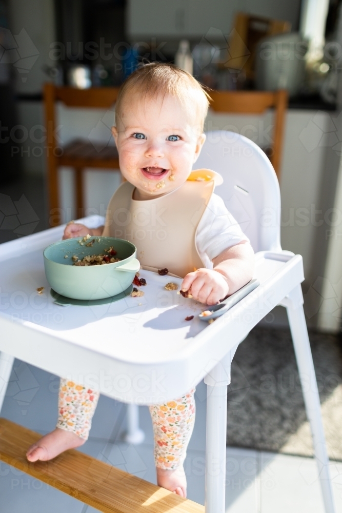 Happy seven month old baby eating breakfast in highchair in morning sunlight - Australian Stock Image