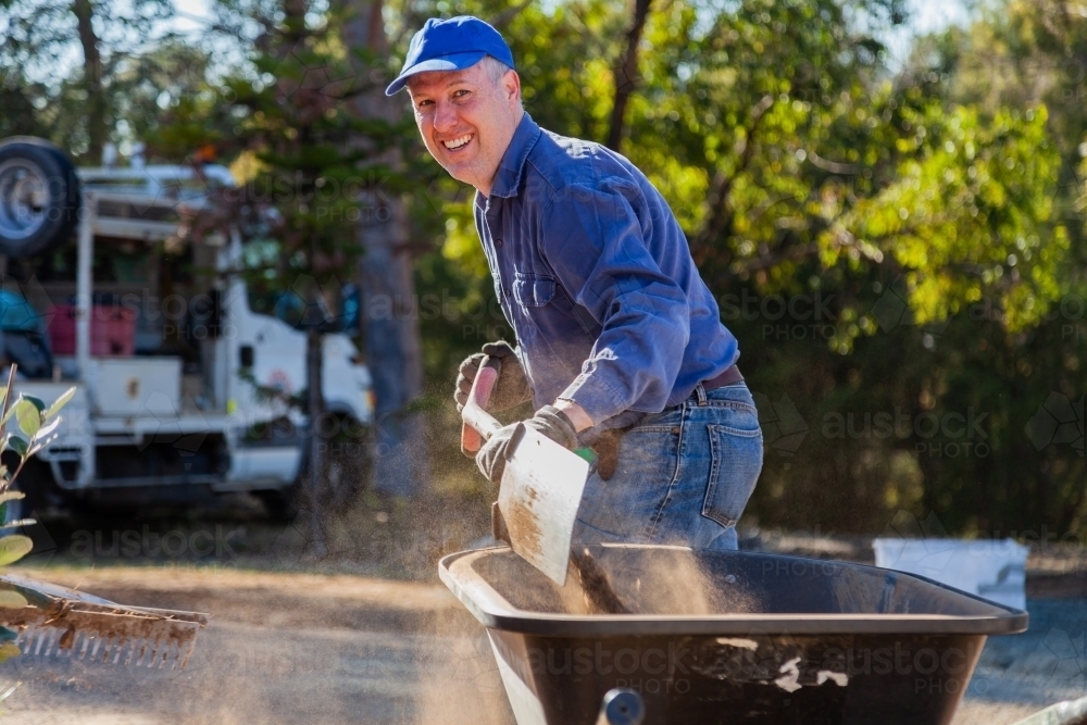 Happy smiling man shoveling dry dusty dirt into a wheelbarrow, gardening - Australian Stock Image