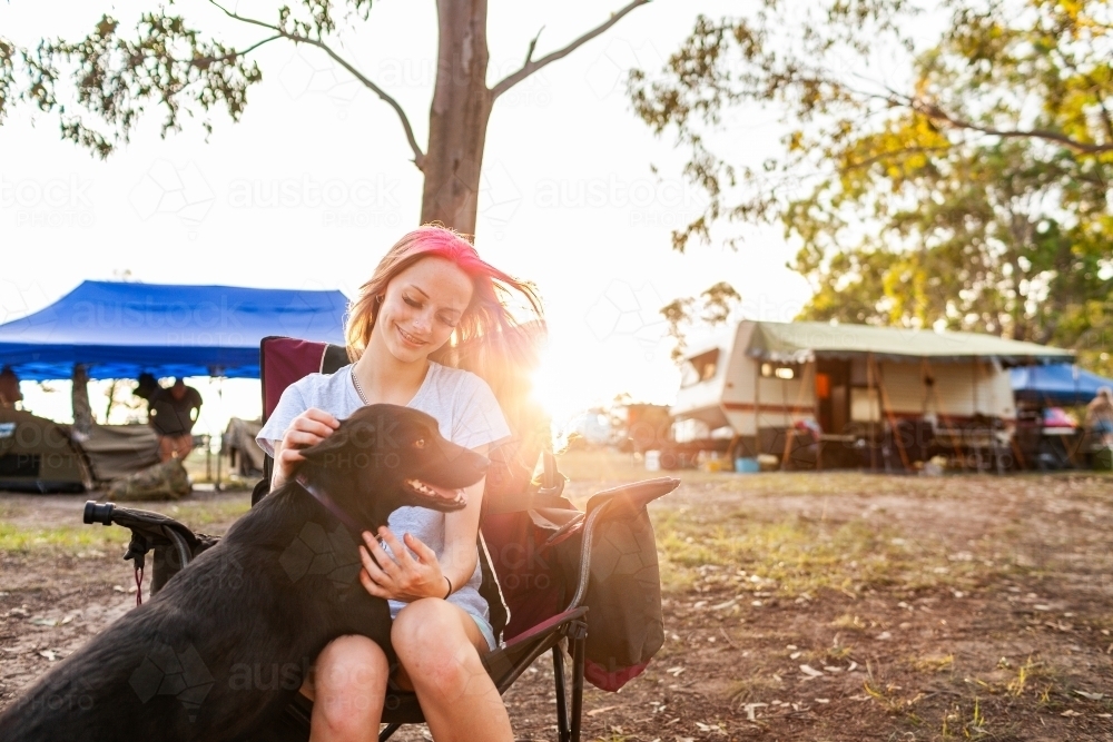 Happy teen girl sitting in camp chair at campsite with her dog on the weekend - Australian Stock Image