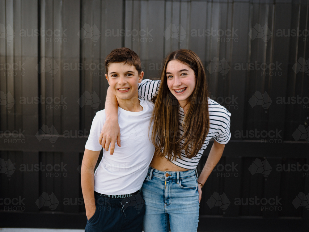 happy teen siblings standing outside against black garage door background - Australian Stock Image