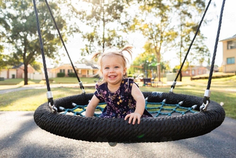 Happy toddler girl on swing at park with playground in backdrop - Australian Stock Image