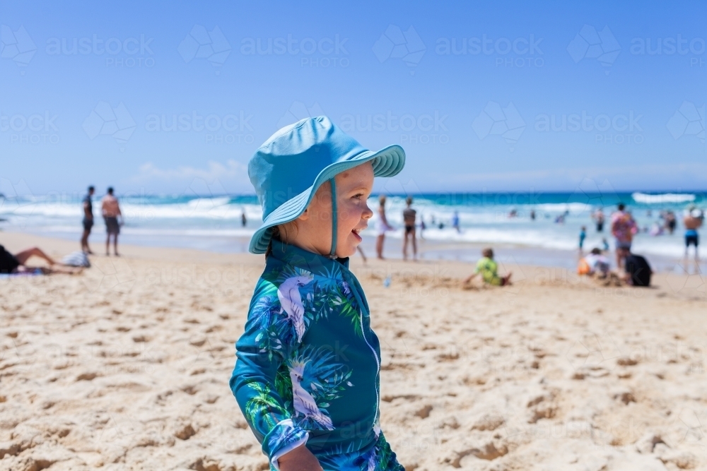 Happy toddler wearing hat for sun protection at beach playing in sand - Australian Stock Image