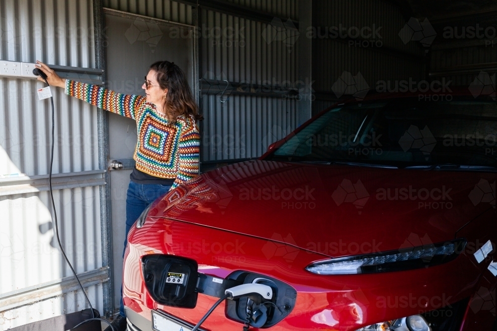 Happy woman in shed garage switching on the electric vehicle car charger at the wall - Australian Stock Image