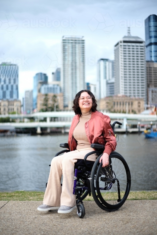 Happy woman in wheelchair in front of river with city in background - Australian Stock Image